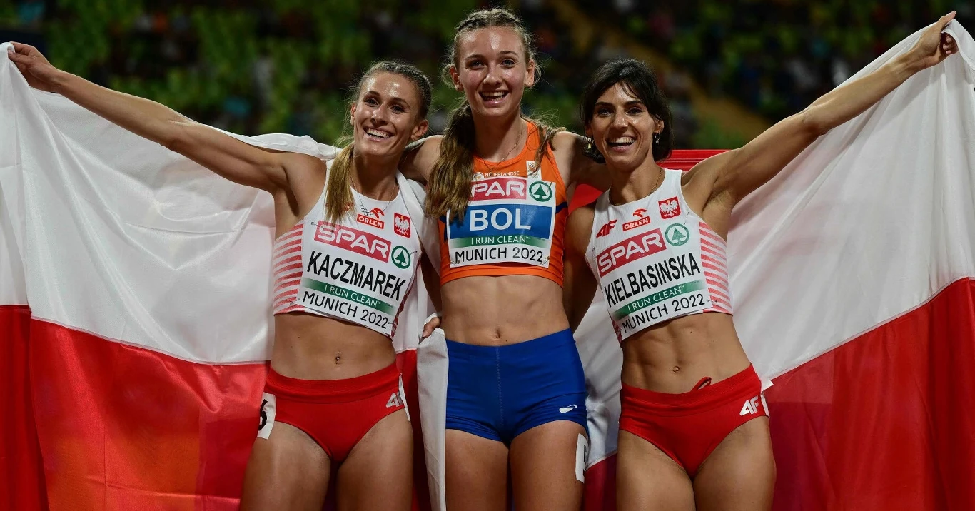 Trzy uśmiechnięte lekkoatletki stoją ramię w ramię na stadionie lekkoatletycznym, trzymając rozpostartą polską flagę. Dwie z nich mają stroje sportowe z polskimi barwami i widocznymi nazwiskami oraz numerami startowymi, a trzecia w środku w niebiesko-p...