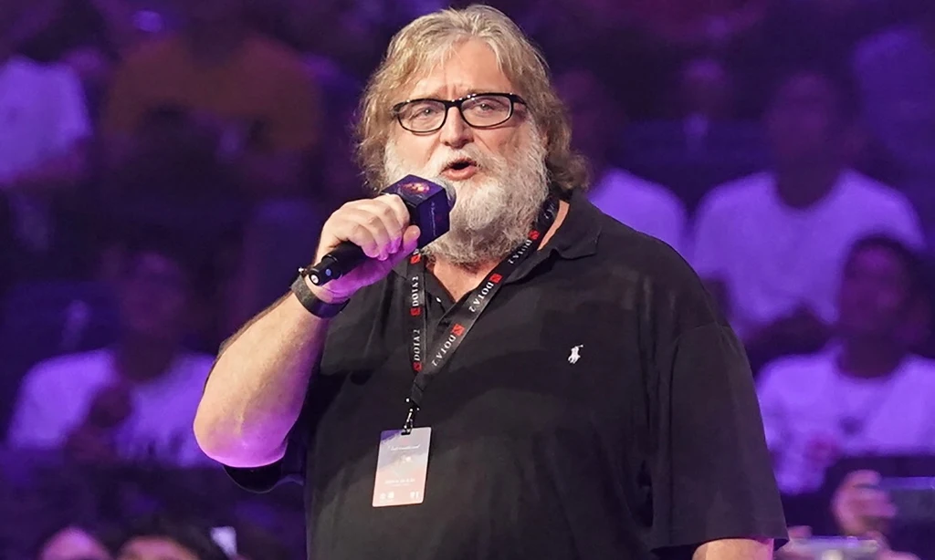 Mężczyzna z mikrofonem w ręku w czarnej koszulce polo, z identyfikatorem na szyi, stoi na scenie podczas wydarzenia publicznego, w tle widownia siedząca w półmroku.