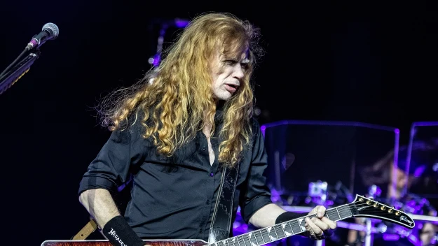 Muzyk z długimi rudymi włosami gra na gitarze elektrycznej na scenie koncertowej, ubrany w czarną koszulę, na nadgarstku widoczna opaska, w tle elementy perkusji i oświetlenie sceniczne.