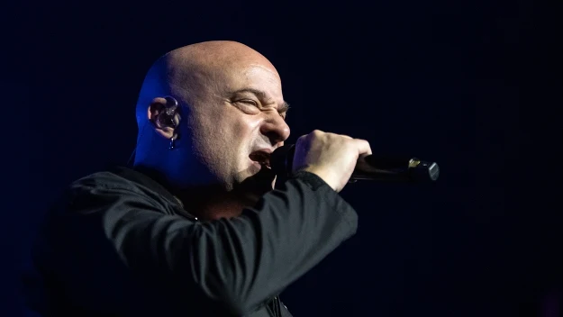 Mężczyzna śpiewający do mikrofonu na ciemnym tle sceny, widoczne emocje podczas występu muzycznego, wyrazista mimika i dynamiczna poza są centralnymi elementami kadru.