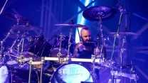 Perkusista grający na rozbudowanym zestawie perkusyjnym podczas koncertu, dynamiczne oświetlenie sceniczne w odcieniach fioletu i niebieskiego, widoczne logo zespołu na bębnie basowym.