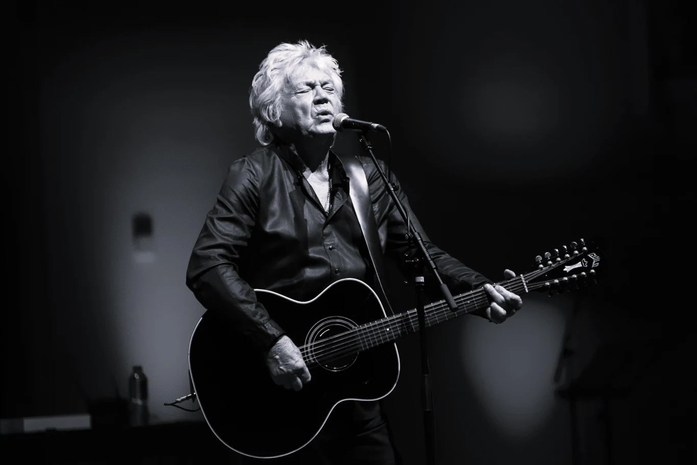 John Lodge z The Moody Blues nie żyje Starszy mężczyzna z siwymi włosami śpiewa do mikrofonu, trzymając czarną gitarę akustyczną, scena oświetlona punktowym światłem, tło rozmyte i ciemne.