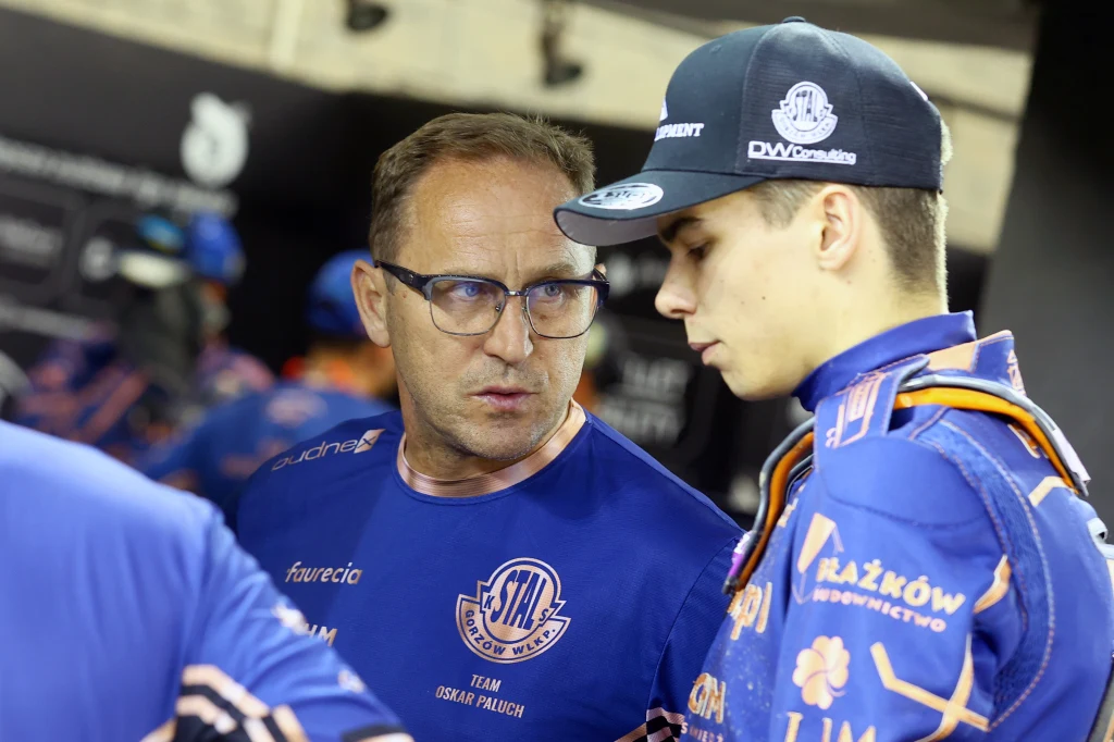 Piotr Paluch z synem Oskarem. Dwóch mężczyzn ubranych w niebieskie stroje drużynowe prowadzi rozmowę. Jeden z nich nosi okulary i patrzy na rozmówcę, który jest młodszy i ubrany w sportowy strój z logo klubu oraz czapkę. W tle rozmyty widok zaplecza sportowego.