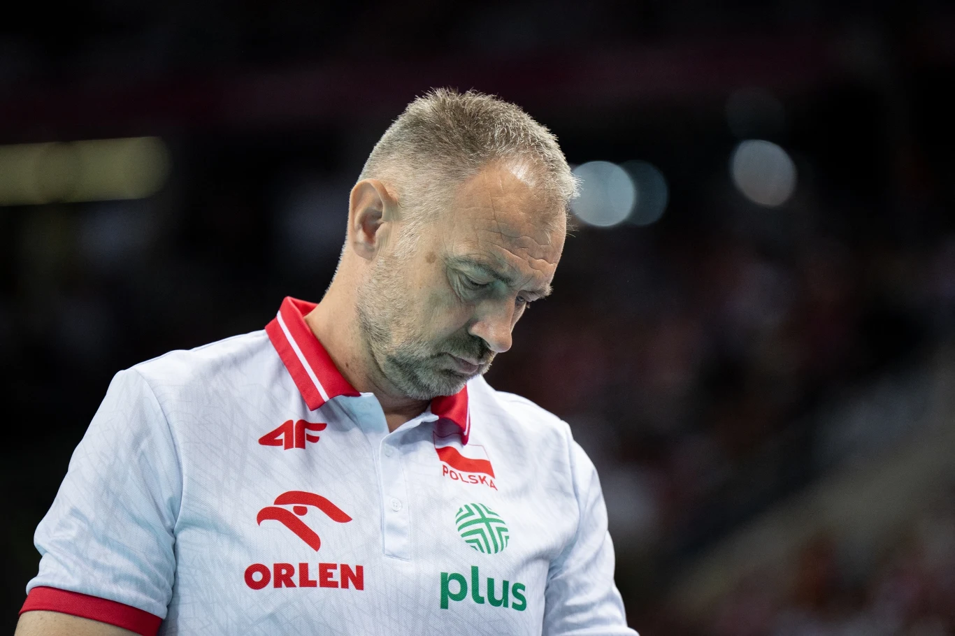 Nikola Grbić Trener polskiej reprezentacji siatkówki w białej koszulce z logotypami sponsorów pochylony w zamyśleniu podczas meczu, z rozmytym tłem hali sportowej.