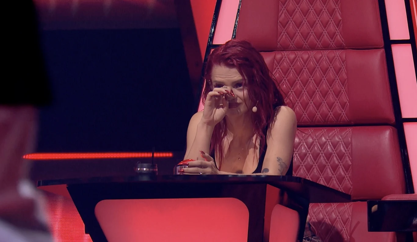 Margaret zalała się łzami po występie jednego z duetów w Bitwach "The Voice of Poland" Kobieta z rudymi włosami siedzi przy dużym panelu na czerwonym, pikowanym fotelu i ociera łzę, wskazując na silne emocje. Przed nią znajduje się mikrofon nagłowny, scena oświetlona jest intensywnym czerwonym światłem.