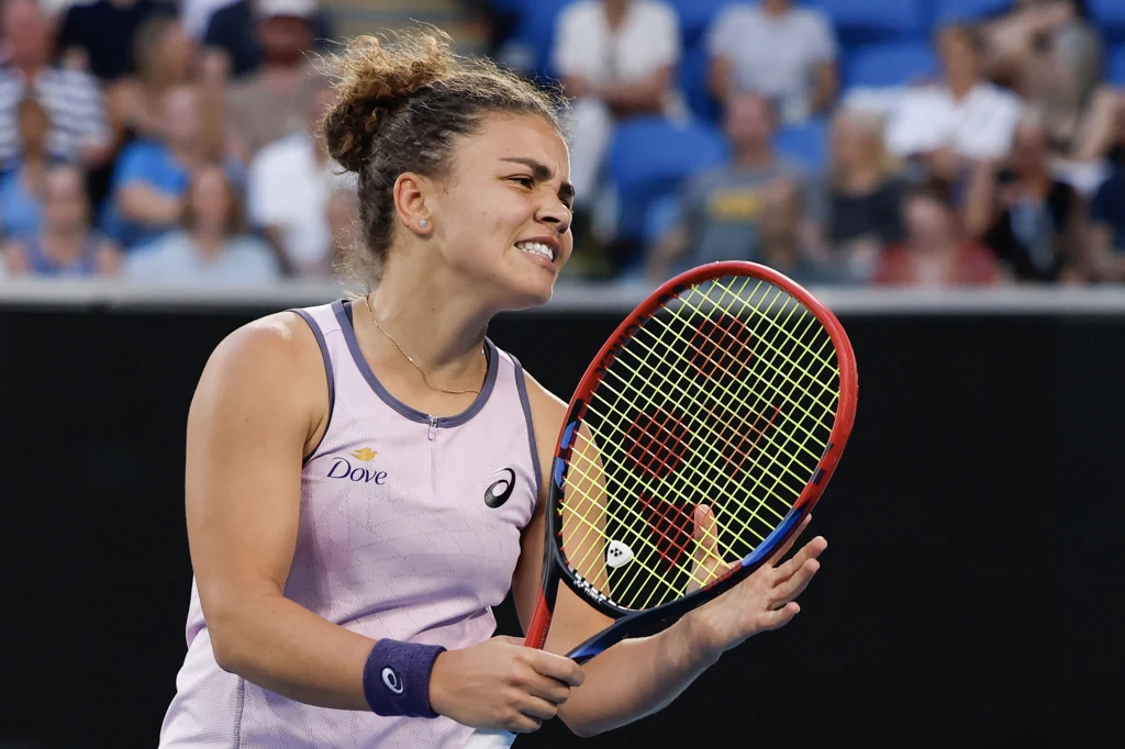 Tenisistka ubrana w fioletową koszulkę i opaskę na nadgarstku trzyma rakietę tenisową i wyraża emocje podczas gry na korcie, w tle widać rozmytych kibiców na trybunach.