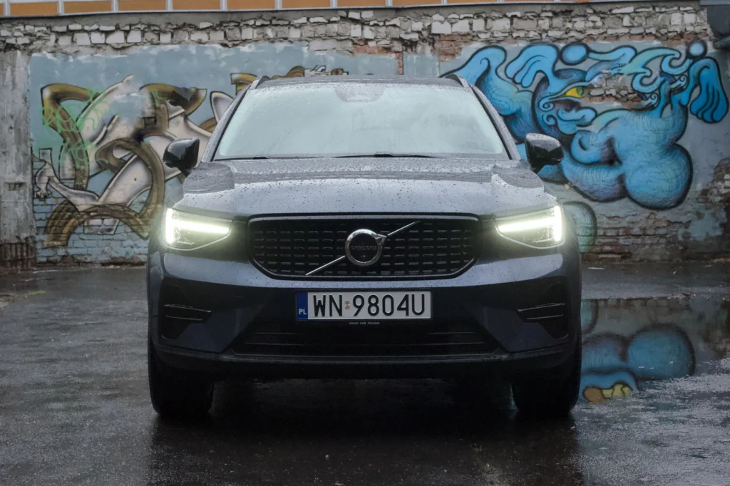 Volvo XC40 mimo upływu lat wciąż wygląda świeżo. Model co prawda przeszedł modernizację w 2022 roku, jednak pod kątem wyglądu zewnętrznego była ona konsmetyczna. Czarny samochód marki Volvo z włączonymi światłami mijania stoi na mokrym asfalcie na tle ściany pokrytej kolorowym graffiti i zniszczonym murem.