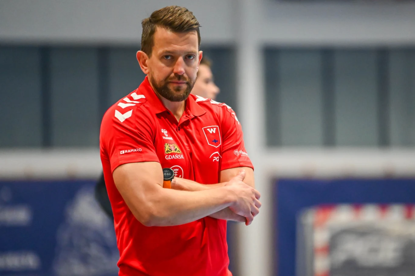 Patryk Rombel Mężczyzna w czerwonej koszulce sportowej z logotypami sponsorów oraz klubem, z założonymi rękami, stoi w hali sportowej i patrzy przed siebie