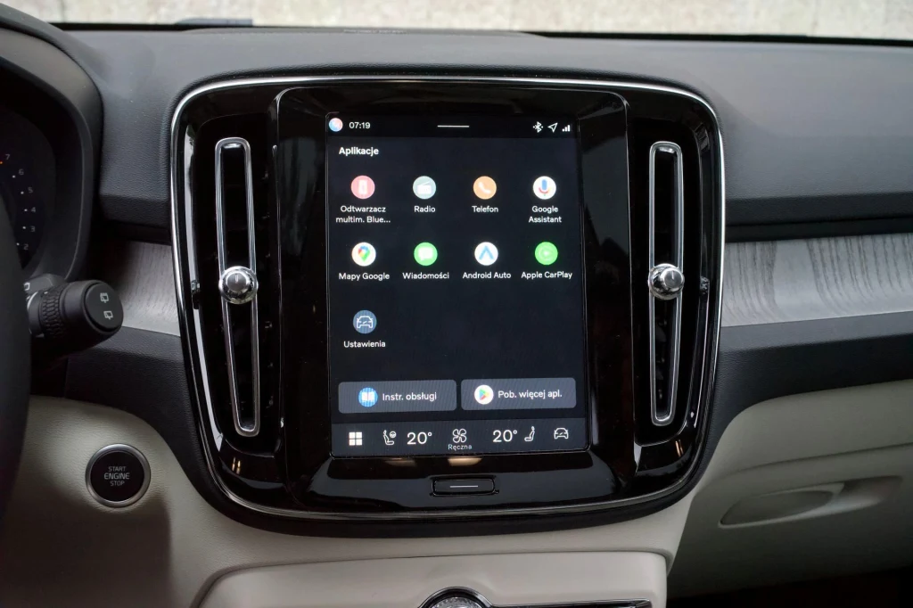 System multimedialny Volvo XC40 wyposażony jest we wbudowane usługi Google, w tym Mapy Google. Aplikacje można pobierać za pośrednictwem Sklepu Play. Centralny ekran dotykowy systemu multimedialnego samochodu z wyświetlonym interfejsem Android Auto, otoczony czarną ramką i pionowymi nawiewami, poniżej przyciski klimatyzacji i panel sterowania.