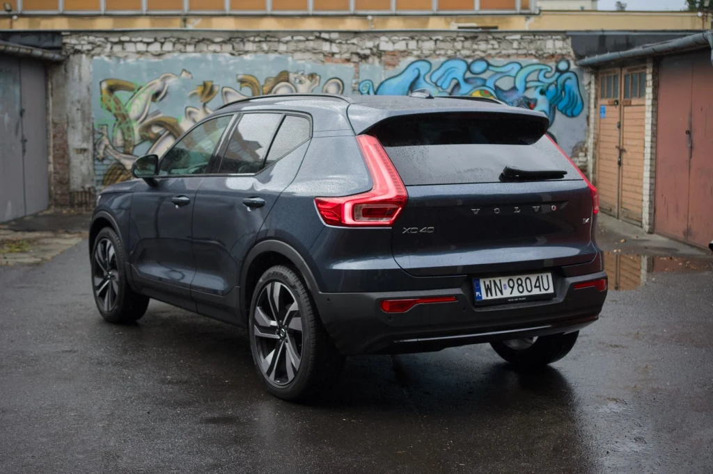 Volvo XC40 dostępne jest obecnie z dwoma wariantami miękkiej hybrydy. W obu bazą jest dwulitrowy silnik benzynowy. Samochód Volvo XC40 w kolorze grafitowym zaparkowany na mokrym asfalcie przed rzędem garaży, w tle ściana z graffiti.