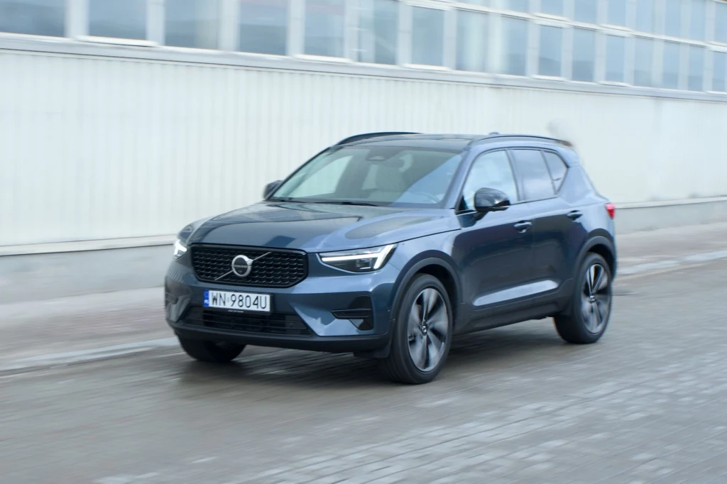 197-konny silnik benzynowy wspierany przez 14-konny silnik elektryczny zapewnia dobrą dynamikę. Nowoczesny, niebieski SUV marki Volvo poruszający się po asfaltowej drodze na tle jasnego, industrialnego budynku.