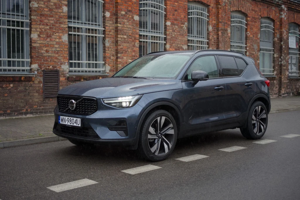 Volvo XC40 kosztuje dziś co najmniej 176 900 zł. Nowoczesny SUV Volvo XC40 w kolorze niebieskim zaparkowany przy zabytkowym, ceglanym budynku z dużymi oknami zabezpieczonymi kratami, asfaltowa ulica z oznaczeniem przejścia dla pieszych.