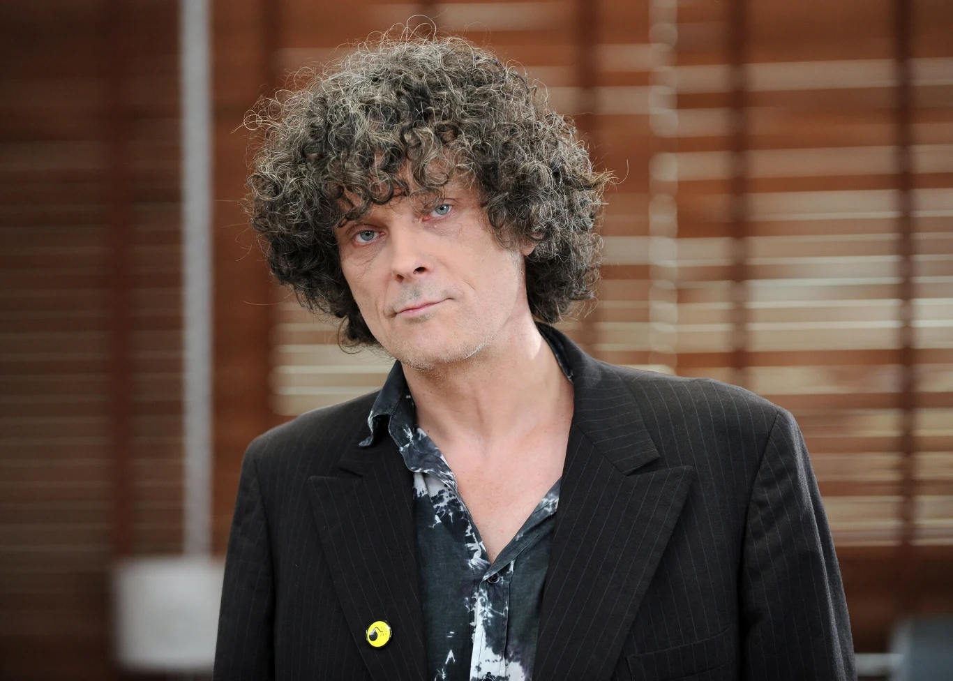 Paweł "Kelner" Rozwadowski był legendą polskiej sceny niezależnej Mężczyzna z kręconymi włosami ubrany w ciemną, prążkowaną marynarkę oraz koszulę z abstrakcyjnym wzorem, stojący na tle brązowych żaluzji, z neutralnym wyrazem twarzy.