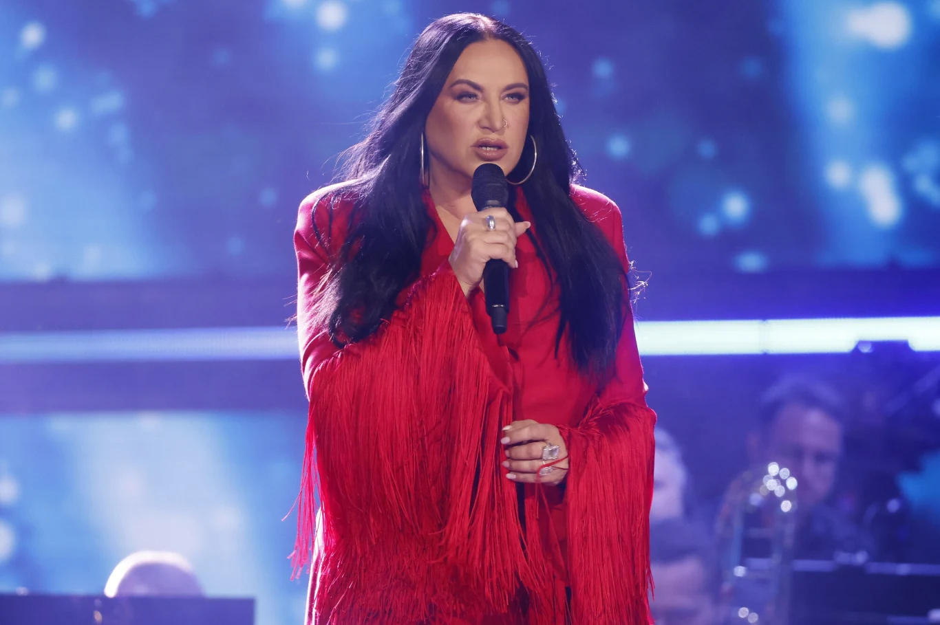 Kayah podziękowała fanom za wsparcie Kobieta w długim czerwonym, frędzlowanym stroju śpiewa na scenie, trzymając mikrofon, w tle rozmyte światła i muzycy.