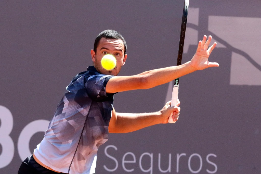 Mężczyzna ubrany w sportową koszulkę odbija piłkę tenisową rakietą na korcie, koncentrując się na precyzyjnym uderzeniu.