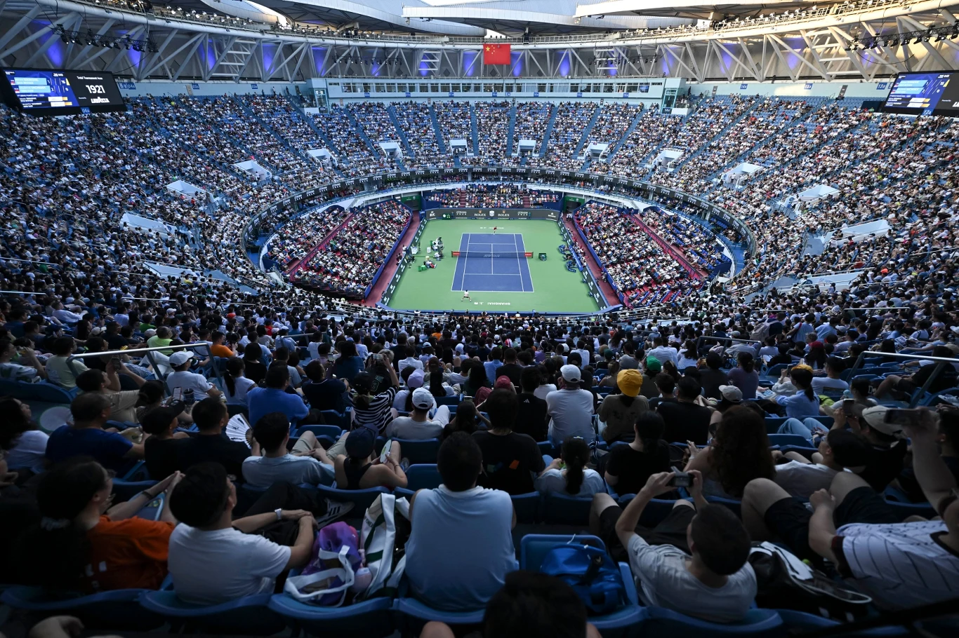 Tłum kibiców zgromadzony na dużym stadionie podczas meczu tenisa, widoczny kort tenisowy na środku, otoczony trybunami wypełnionymi widzami.