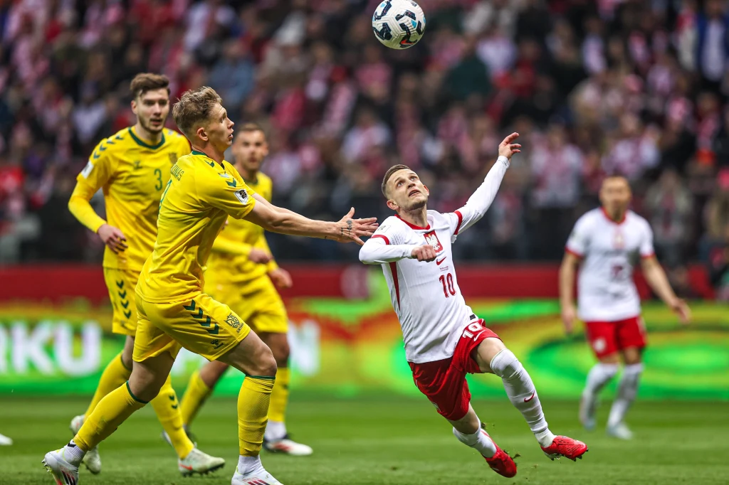 Dwóch piłkarzy w dynamicznej walce o piłkę podczas meczu, jeden z zawodników w białej koszulce próbuje przechwycić piłkę w powietrzu, otoczony przez rywala w żółtym stroju, w tle znajduje się tłum kibiców.