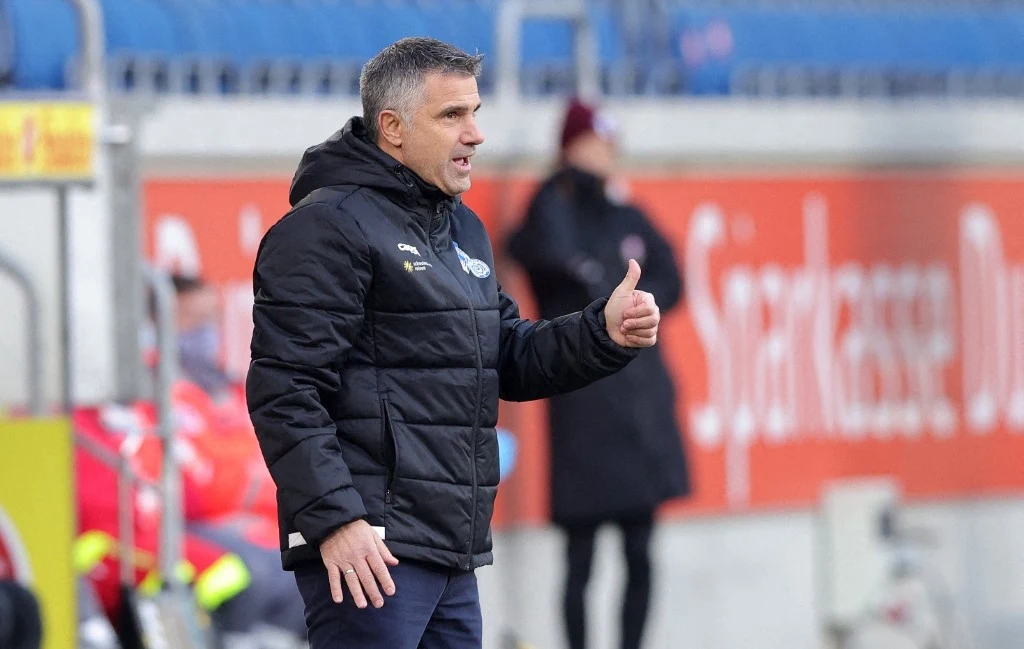 Gino Lettieri zadebiutował na ławce Wieczystej Kraków Mężczyzna w czarnej kurtce sportowej robi gest kciuka w górę, stojąc przy linii bocznej boiska, w tle rozmyte postacie i reklamy.