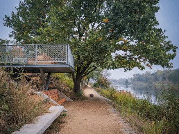 Szeroka, piaszczysta ścieżka biegnąca wzdłuż rzeki, z ławkami i tarasem widokowym wspartym na metalowej konstrukcji, otoczona zielenią i dużym drzewem w cieniu którego można odpocząć.