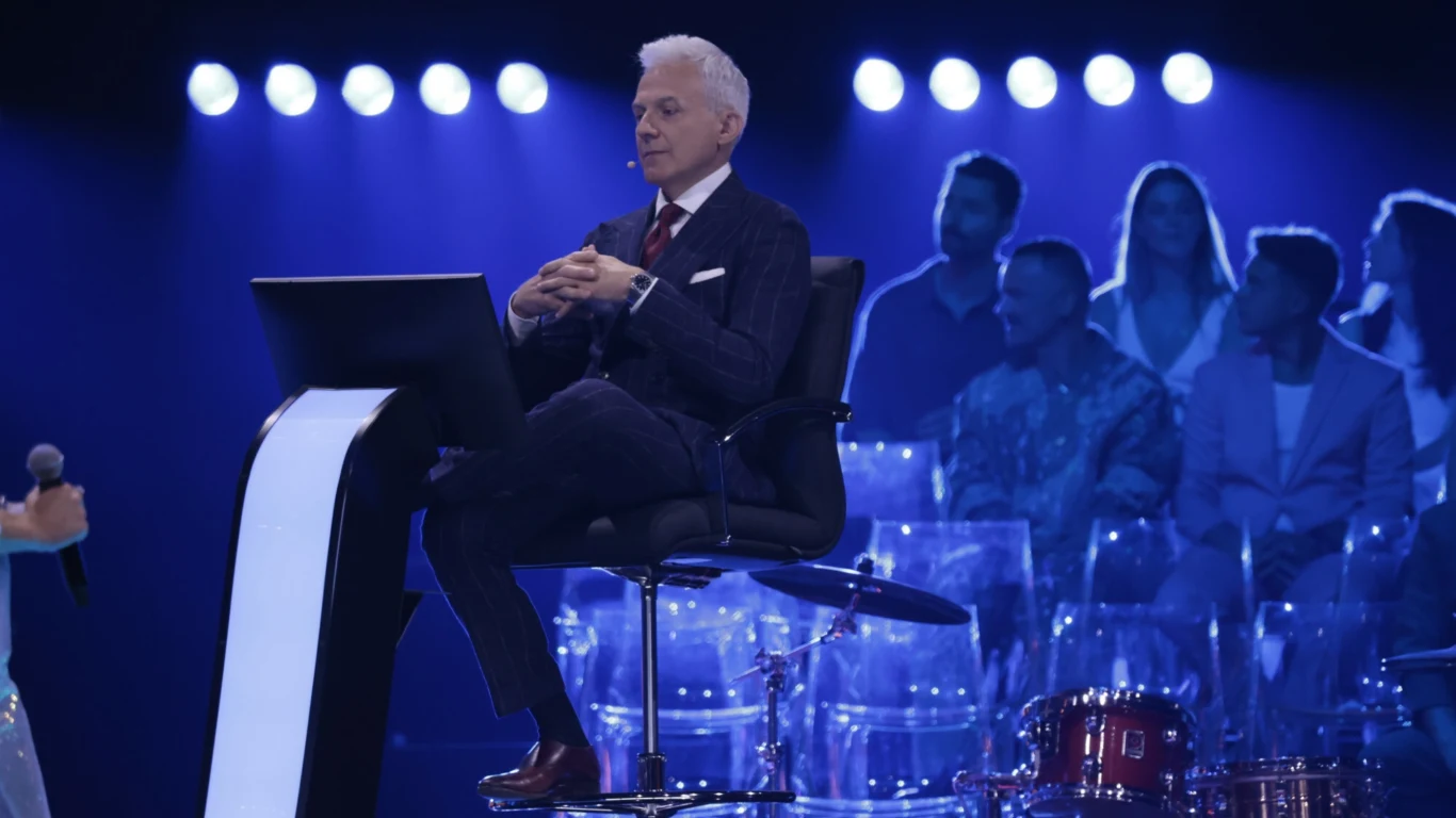 Milionerzy Elegancko ubrany mężczyzna siedzi na wysokim, nowoczesnym krześle przy podświetlonym pulpicie, w tle widać grupę ludzi na przezroczystych siedzeniach oraz fragment zestawu perkusyjnego, całość oświetlona niebieskim światłem.