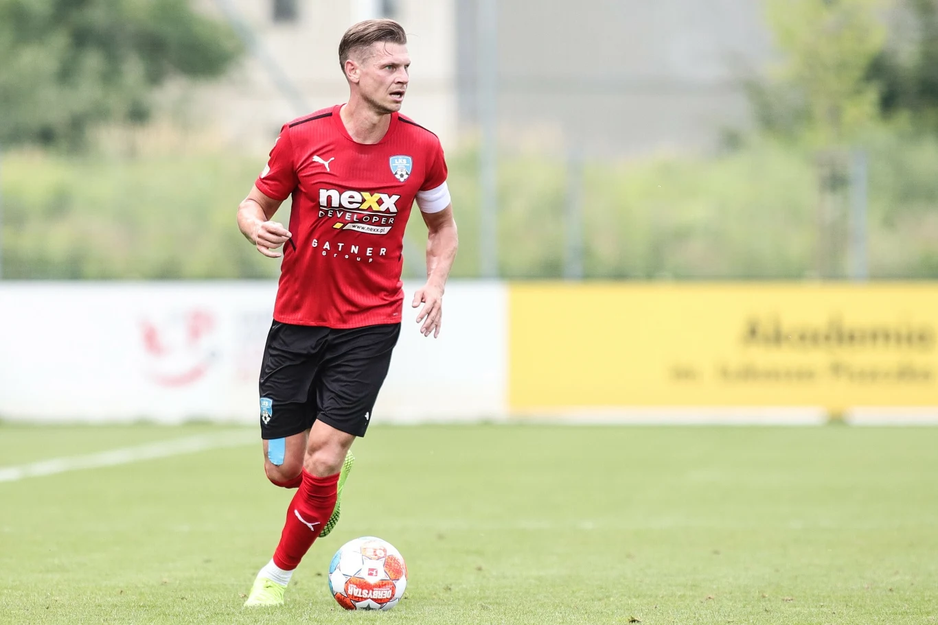 Łukasz Piszczek broni barw LKS Goczałkowice - Zdrój Piłkarz w czerwonej koszulce i czerwonych getrach prowadzi piłkę na zielonym boisku podczas meczu, w tle rozmyte elementy infrastruktury sportowej i banery reklamowe.