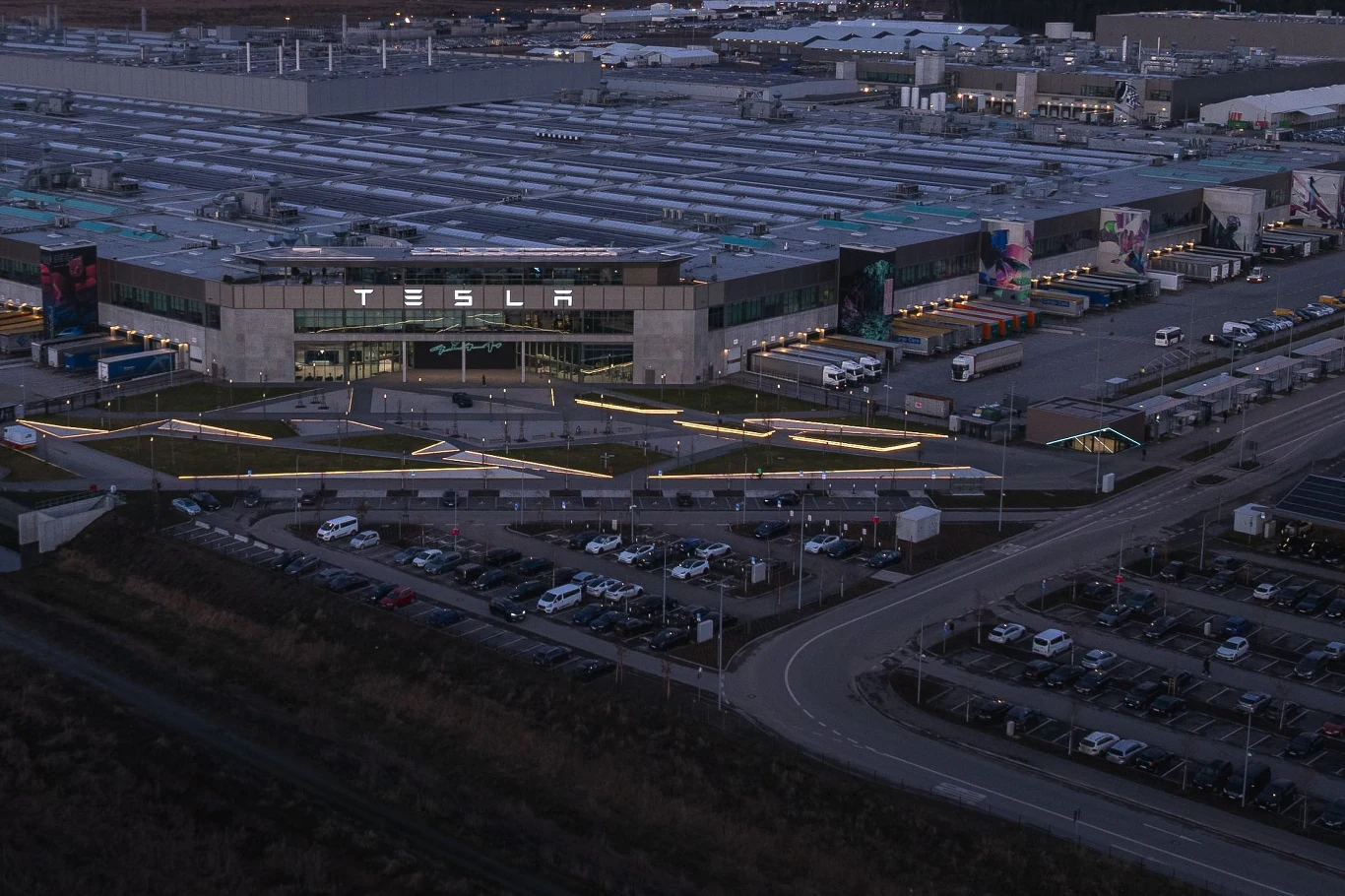 Fabryka samochodów elektrycznych Tesla z dużym napisem na głównym budynku, otoczona licznymi samochodami na parkingu oraz ciężarówkami dostawczymi, nowoczesny kompleks przemysłowy z widocznymi liniami dojazdowymi i infrastrukturalnymi.