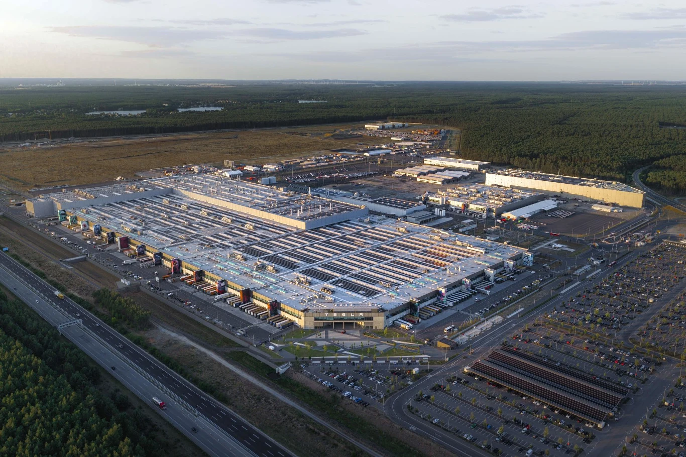 Ogromny kompleks przemysłowy otoczony rozległymi parkingami oraz terenami leśnymi, szeroka droga szybkiego ruchu biegnie wzdłuż jednej z granic obiektu, wokół widoczne są liczne magazyny oraz hale logistyczne.