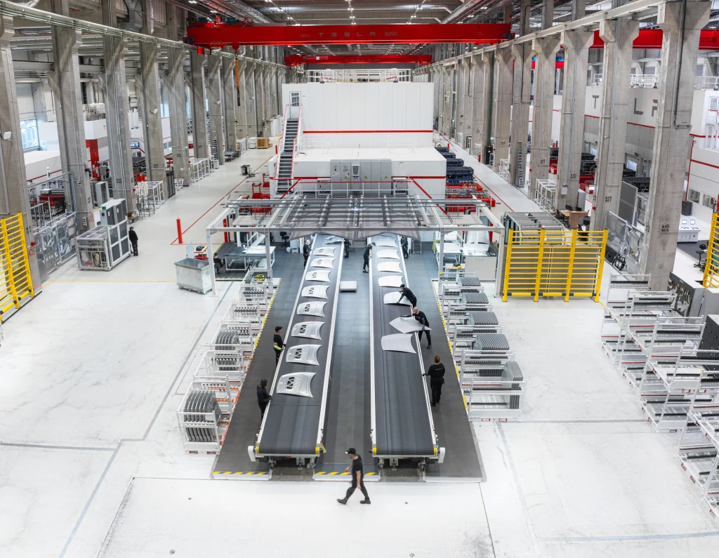 Również elementy karoserii powstają tu na miejscu – wyciskane przez wielkie prasy z blach składowanej w rolkach Nowoczesna hala produkcyjna z wysokim sufitem, rzędy maszyn i taśm produkcyjnych w centrum kadru, kilku pracowników ubranych w ciemne ubrania pracujących przy linii montażowej, liczne regały i żółte barierki ochronne po bokach, czyste i uporządkowane o...