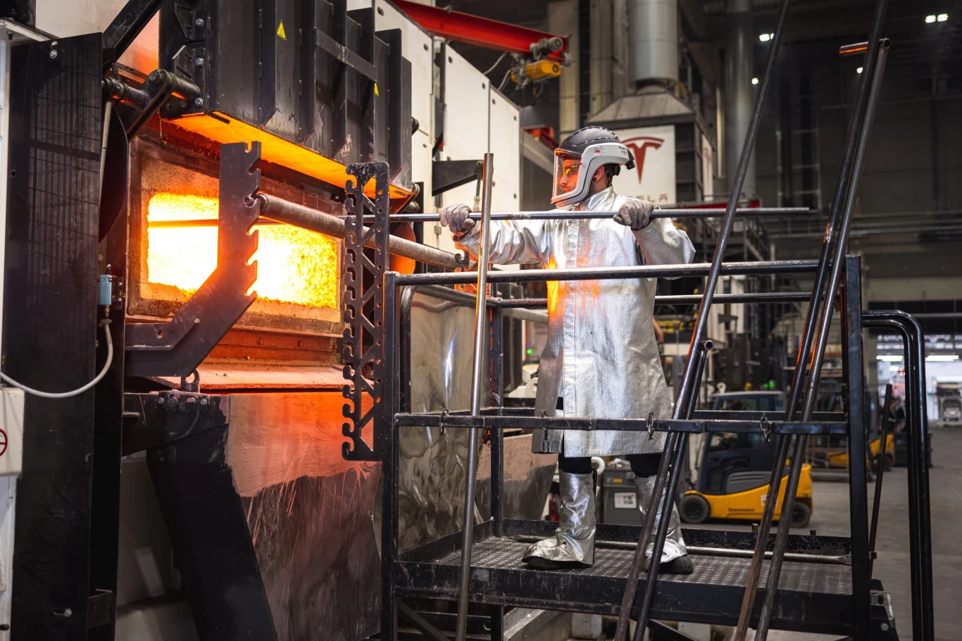 Pracownik ubran w specjalistyczny strój ochronny z przyłbicą obsługuje rozgrzany do wysokiej temperatury piec hutniczy, wykonując swoje obowiązki w przemysłowej hali produkcyjnej.