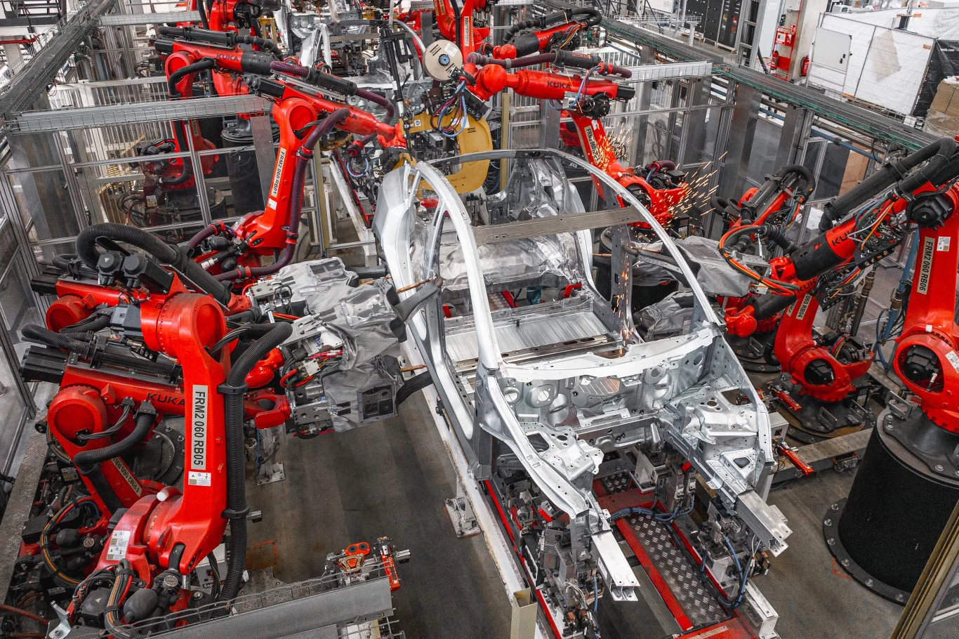 Karoseria samochodu w trakcie montażu przez liczne czerwone roboty przemysłowe umieszczone wokół linii produkcyjnej w nowoczesnej fabryce.