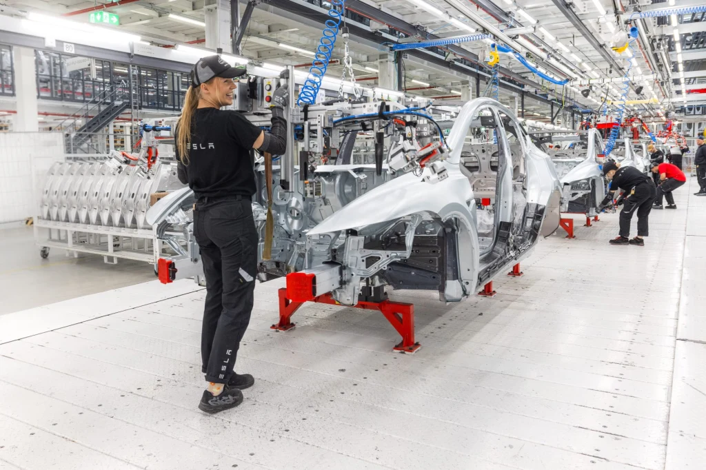 W ciągu dnia w fabryce pod Berlinem powstaje przeszło tysiąc samochodów Pracownica obsługująca maszynę przy linii montażowej w nowoczesnej fabryce samochodów, po lewej widoczny szkielet pojazdu elektrycznego oraz inne osoby pracujące przy kolejnych etapach montażu.