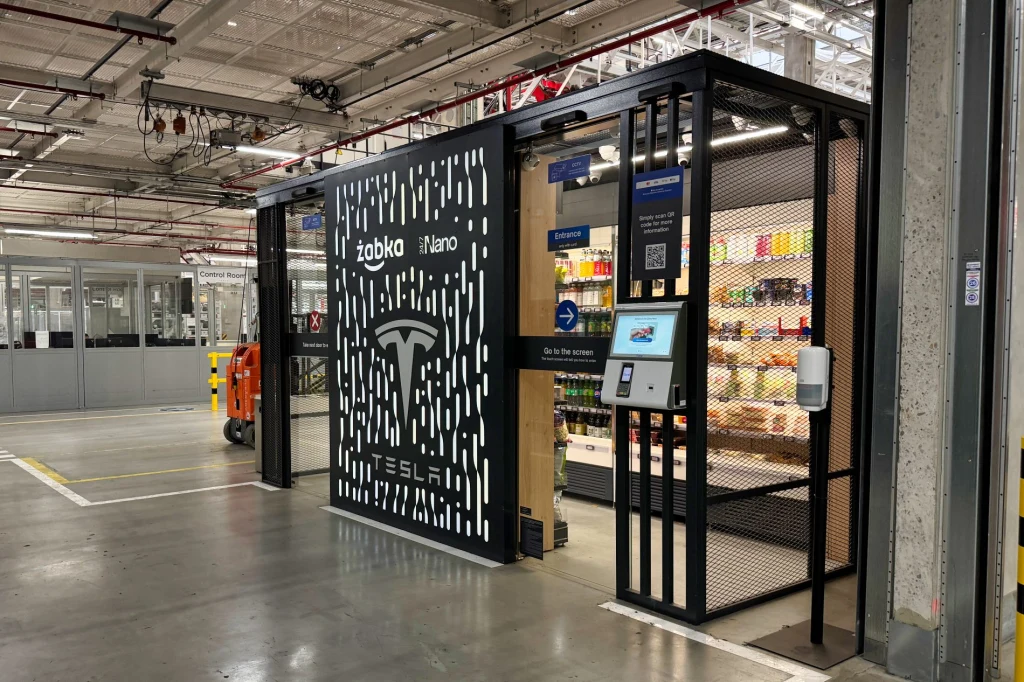 Zarówno w części biurowej jak i na halach produkcyjnych można trafić na Żabki Nano Wejście do automatycznego sklepu Żabka Nano sygnowanego logo Tesli, z widoczną półką sklepową pełną produktów spożywczych oraz elektronicznym panelem dostępowym, całość w środku przemysłowej hali.