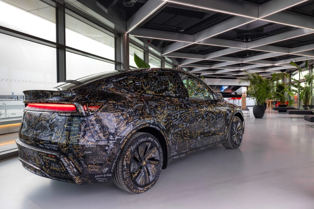 Pierwszy wyprodukowany tu Model Y, ozdobiony podpisami pracowników, stoi w zakładowej kawiarni Nowoczesny samochód elektryczny SUV w futurystycznym wnętrzu salonu, na karoserii liczne grafiki i napisy, geometryczny sufit z oświetleniem LED, duże okna oraz rośliny doniczkowe w tle.