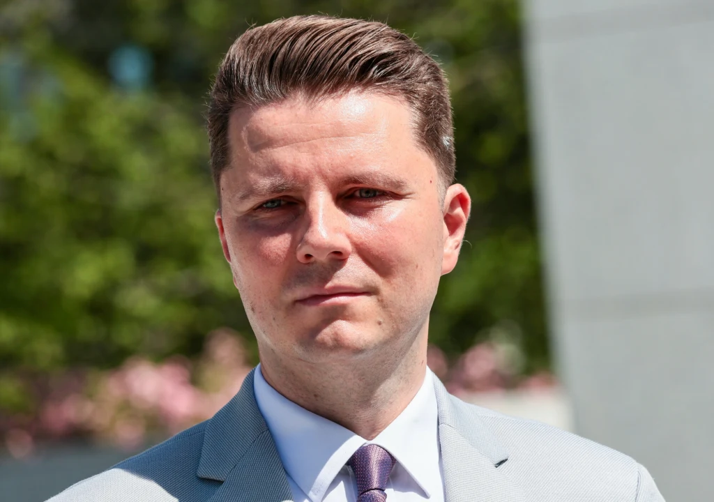 Mężczyzna w garniturze na tle zieleni i niewyraźnych kwiatów, z poważnym wyrazem twarzy, dobrze uczesane włosy, jasna marynarka i krawat, słoneczna pogoda.