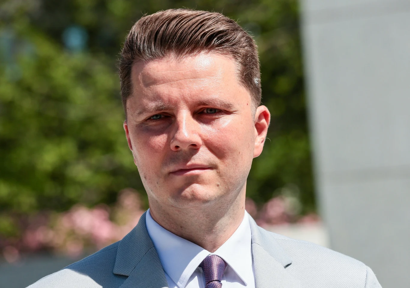 Mężczyzna w garniturze na tle zieleni i niewyraźnych kwiatów, z poważnym wyrazem twarzy, dobrze uczesane włosy, jasna marynarka i krawat, słoneczna pogoda.