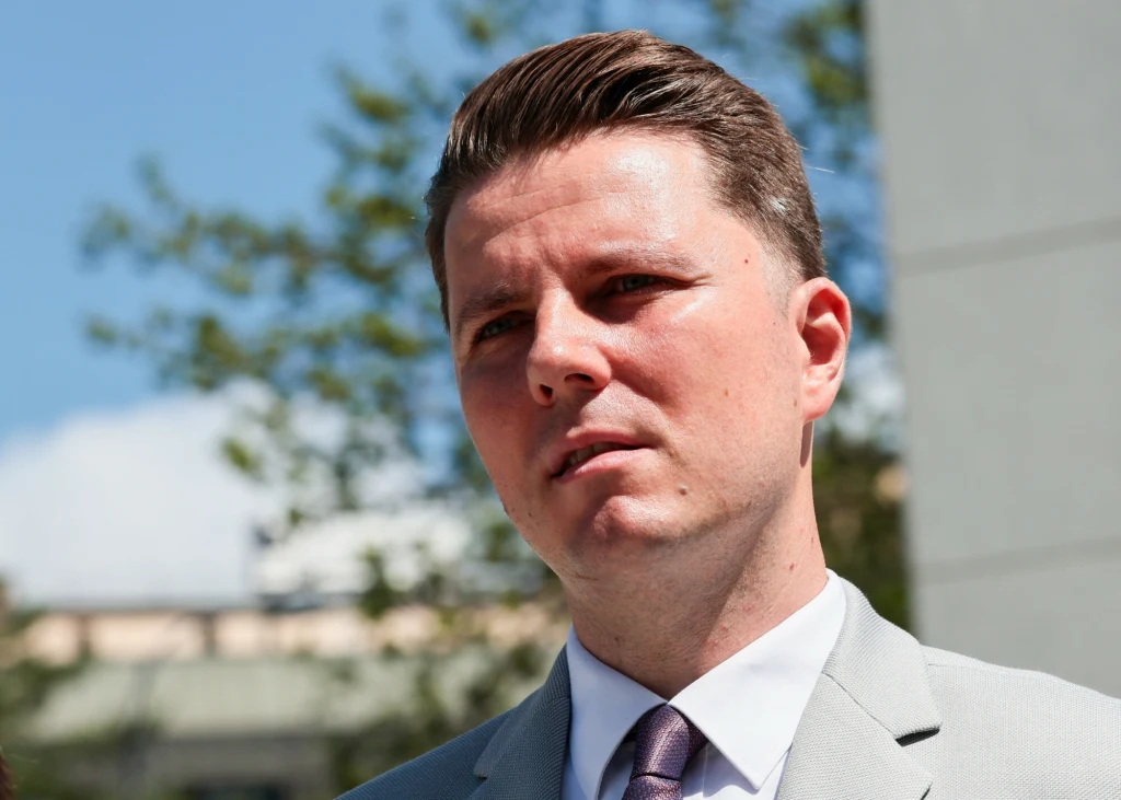 Mężczyzna w jasnym garniturze z poważnym wyrazem twarzy na tle rozmytej zieleni i miejskiego krajobrazu, ujęcie wykonane w świetle dziennym.