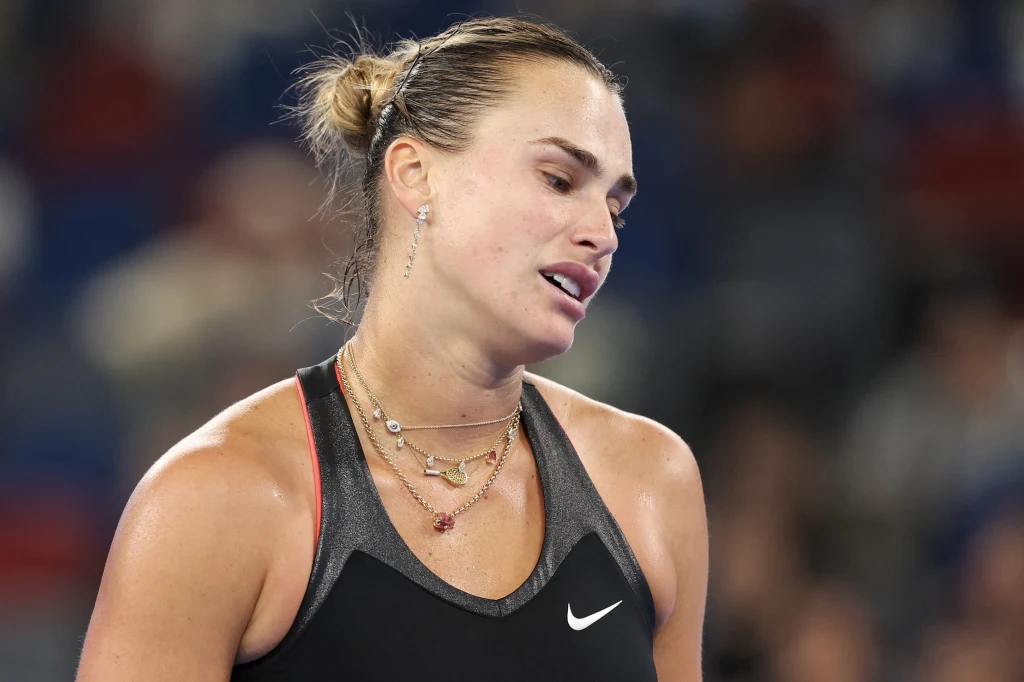 Aryna Sabalenka Kobieta w sportowej, czarnej koszulce z logo Nike, z włosami związanymi w kok, wyrażająca zmęczenie lub rozczarowanie, tło rozmyte, co skupia uwagę na jej twarzy.