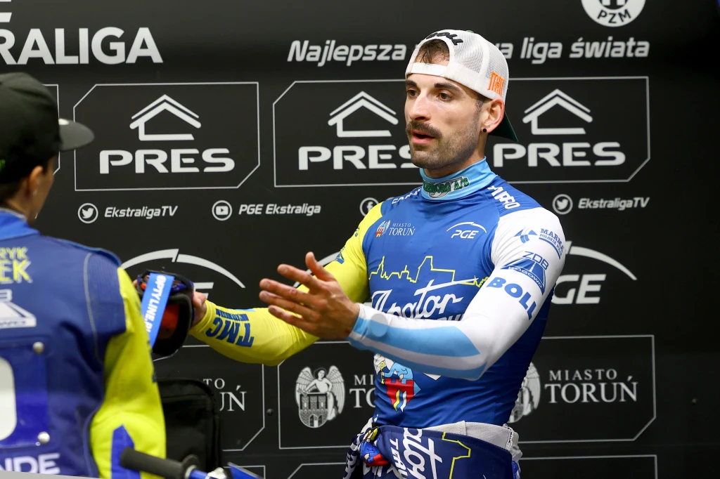 Zawodnik żużlowy w stroju sportowym prowadzi rozmowę z innym uczestnikiem wydarzenia podczas konferencji prasowej lub wywiadu, w tle widoczne są banery sponsorów i logotypy związane z PGE Ekstraligą.