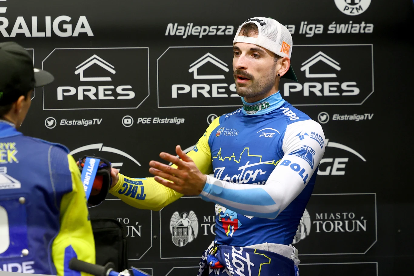 Niezadowolony Mikkel Michelsen. Zawodnik żużlowy w stroju sportowym prowadzi rozmowę z innym uczestnikiem wydarzenia podczas konferencji prasowej lub wywiadu, w tle widoczne są banery sponsorów i logotypy związane z PGE Ekstraligą.