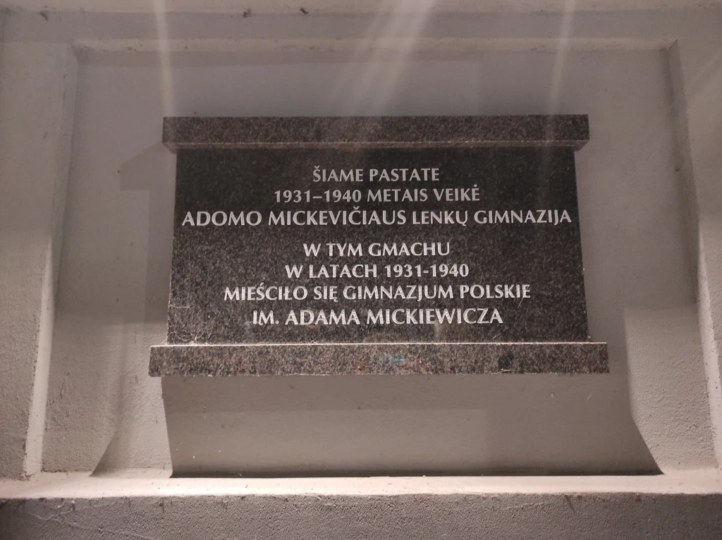Tablica pamiątkowa z czarnego kamienia z wygrawerowanym napisem informującym o działalności polskiego gimnazjum imienia Adama Mickiewicza w latach 1931-1940, zamontowana na jasnej ścianie.