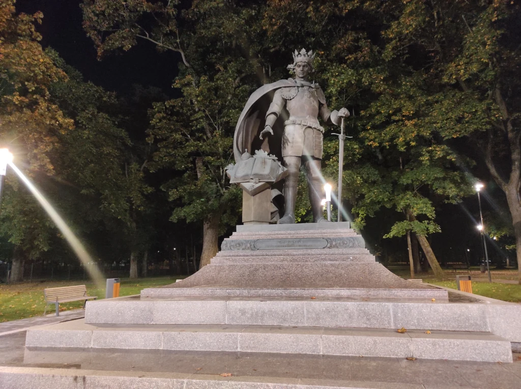 Monumentalny pomnik króla z koroną i mieczem, stojącego na cokole, w tle parkowe drzewa i nocne oświetlenie.