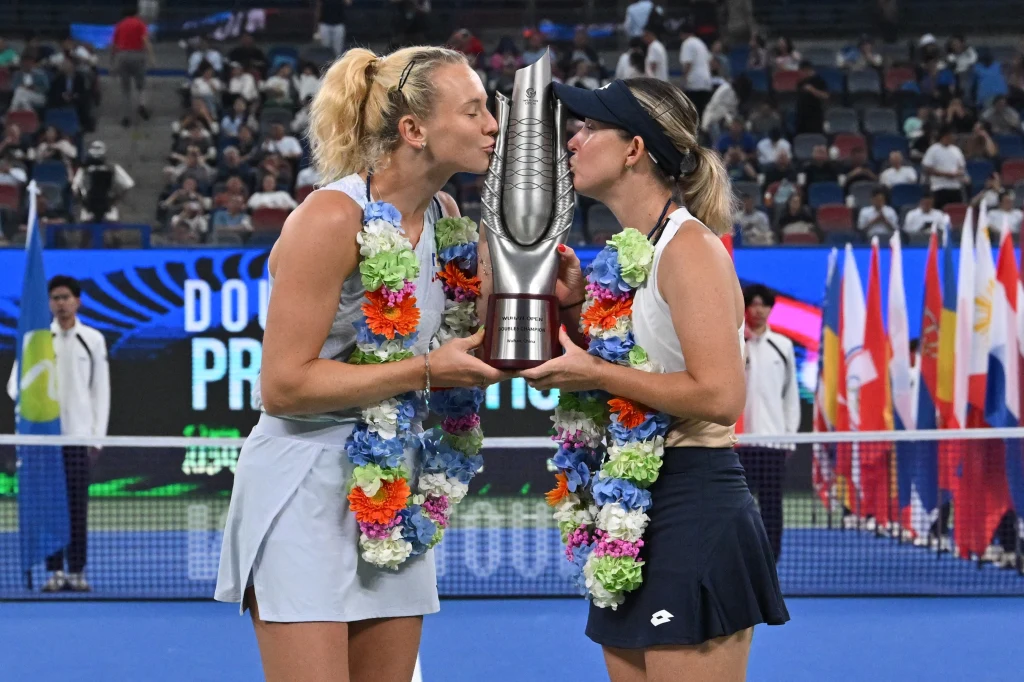 Dwie tenisistki w girlandach kwiatowych całują dużych rozmiarów puchar po zwycięstwie na turnieju tenisowym, w tle kort oraz obserwatorzy i flagi państwowe.