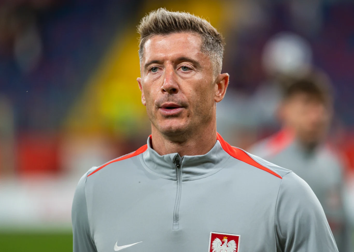 Kapitan reprezentacji Polski - Robert Lewandowski Mężczyzna o jasnych włosach w szarej bluzie sportowej z godłem Polski na piersi, stoi na tle rozmytego stadionu, wyraz twarzy skupiony
