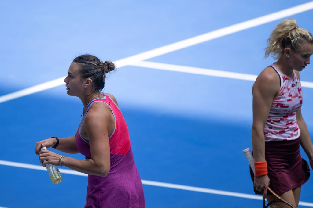 Dwie liderki rankingów WTA: Aryna Sabalenka i Katerina Siniakova. W Wuhanie po tytuł sięgnęła tym razem Czeszka Dwie tenisistki na korcie, jedna trzyma butelkę z napojem, obie ubrane w sportowe stroje, nie wykazujące wyraźnych emocji na twarzach, w tle linie kortu tenisowego.