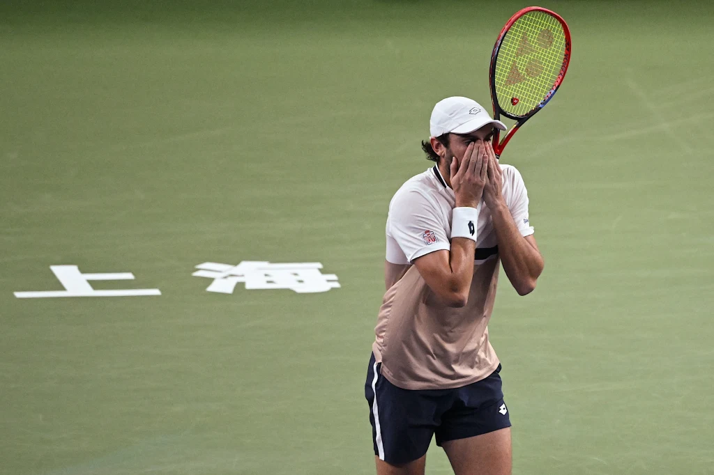Valentin Vacherot Tenisista w jasnej koszulce i białej czapce zakrywa twarz dłońmi, trzymając w drugiej ręce rakietę, stojąc na korcie z chińskim napisem na tle zielonej nawierzchni.