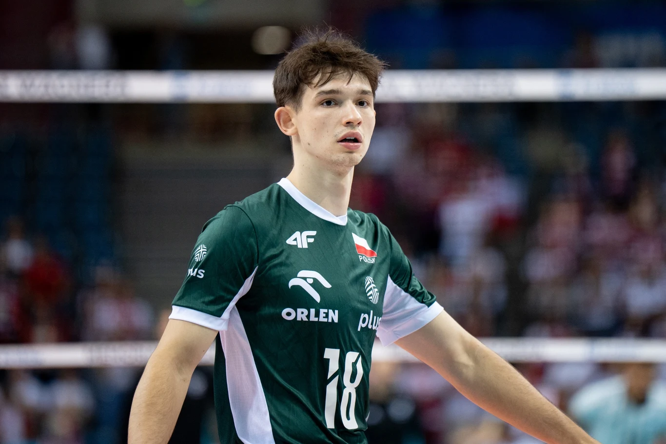 Młody siatkarz w zielonym stroju z numerem 18 na boisku sportowym, w tle rozmyta widownia i hala sportowa.