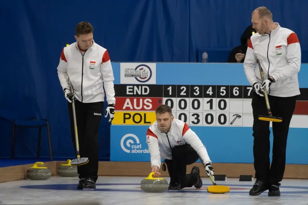 Trzech zawodników drużyny curlingowej w białych kurtkach z czerwonymi elementami rozgrywa mecz, jeden z nich wypuszcza kamień curlingowy po lodzie, w tle widoczna tablica wyników wskazująca na rywalizację drużyn z Polski i Australii.