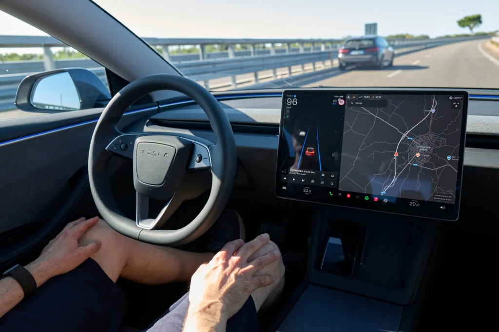 Wnętrze samochodu Tesla podczas jazdy autostradą z widocznym dużym ekranem systemu nawigacji oraz osobą siedzącą na miejscu kierowcy, trzymającą ręce na kolanach, co sugeruje tryb autonomiczny pojazdu.