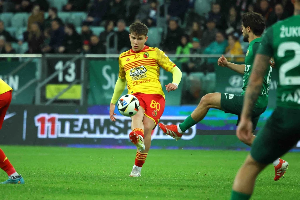 Oskar Pietuszewski Zawodnik w żółto-czerwonym stroju kopie piłkę podczas meczu piłki nożnej, obok niego kilku zawodników w zielonych strojach, a w tle widać trybuny z kibicami.