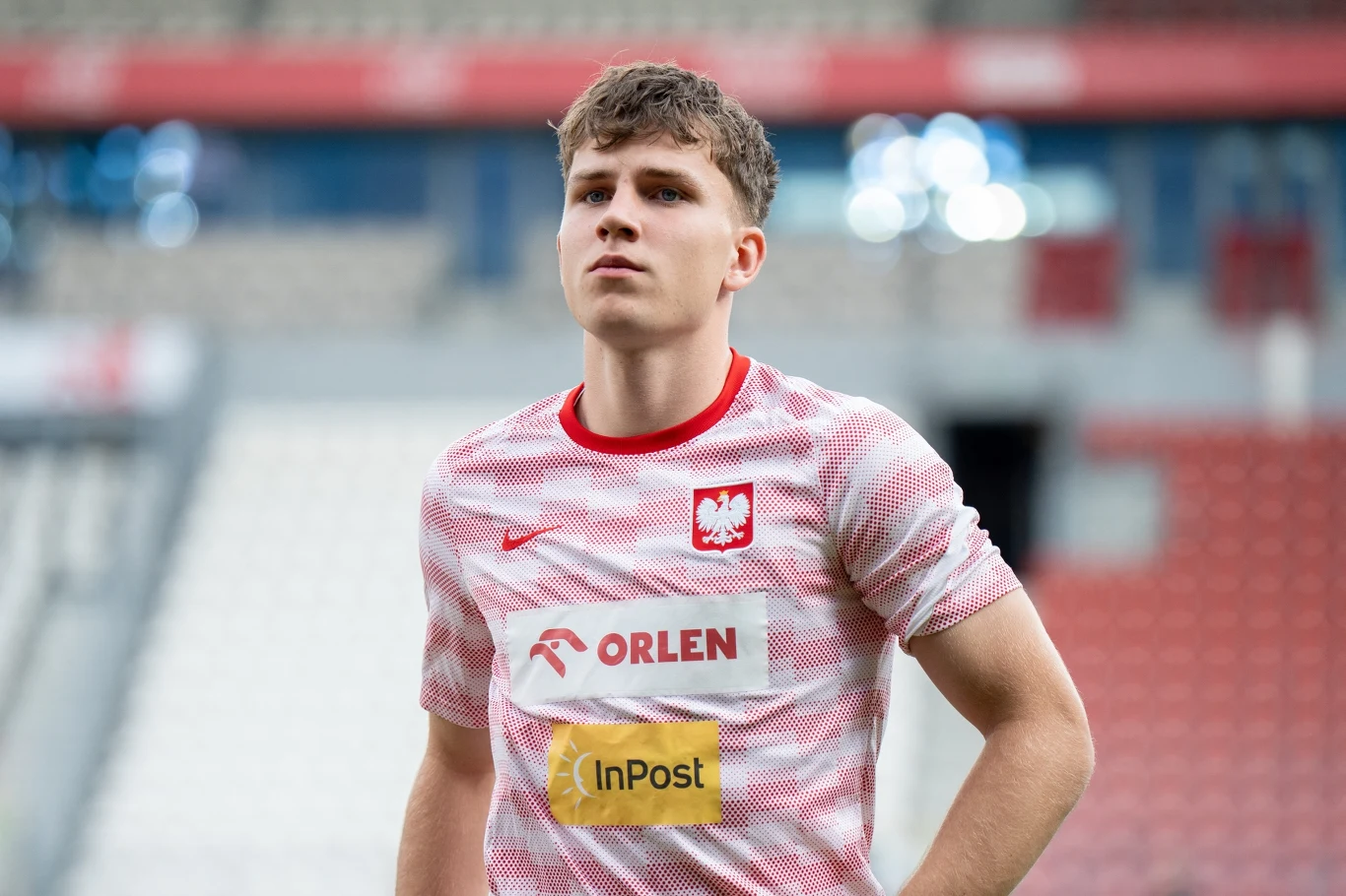 Młody piłkarz w biało-czerwonej koszulce reprezentacji Polski z widocznymi logotypami sponsorów stoi na tle trybun stadionu.